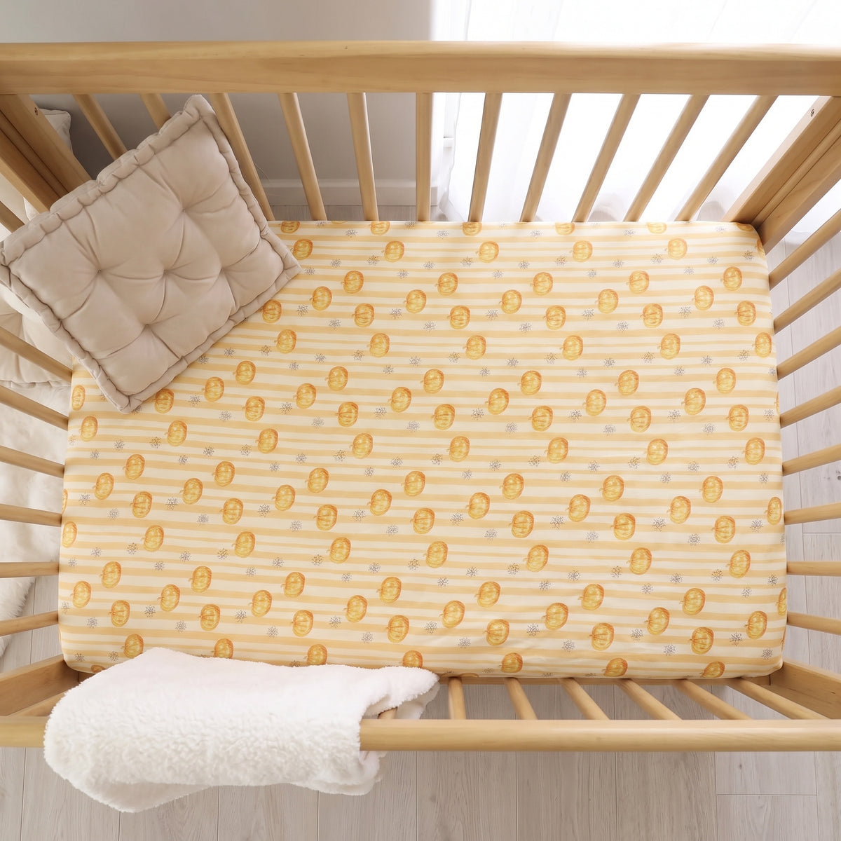 A baby crib with a light wood slat frame, featuring a yellow and white striped mattress cover with a gold coin pattern, a tufted cream pillow, and a white blanket.