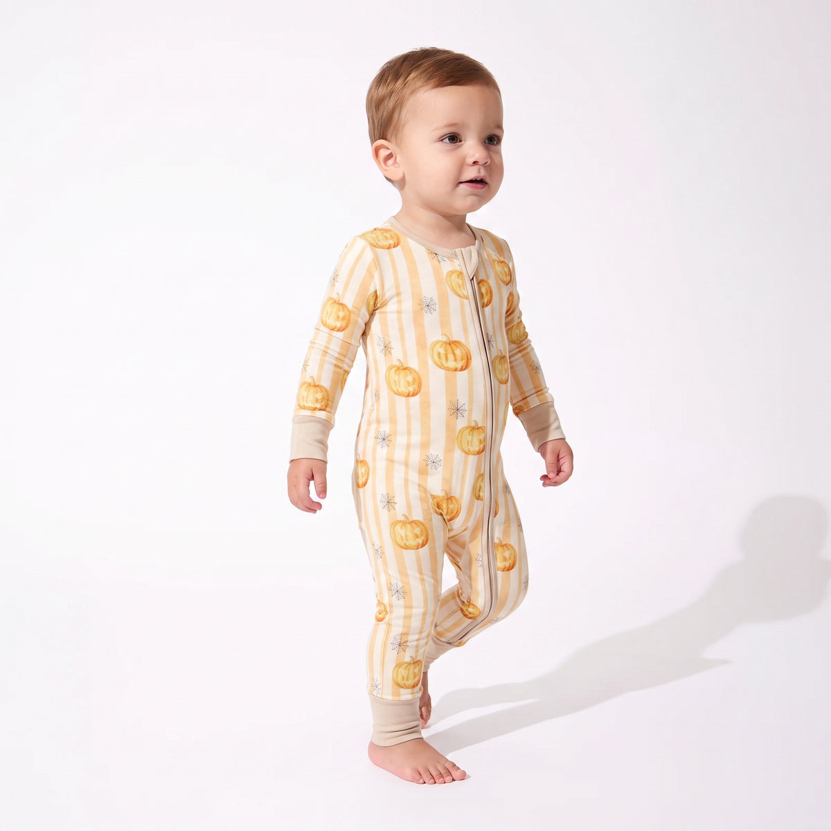 A baby wearing a cream and yellow striped one-piece pajama with a pumpkin print, standing against a white studio background.