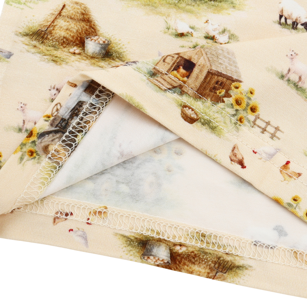 A folded piece of cream-colored fabric printed with a rustic farm scene, featuring a small wooden barn, haystack, chickens, sheep, and sunflowers. The fabric has a decorative white lace-like stitched border and is shown layered on a white background.