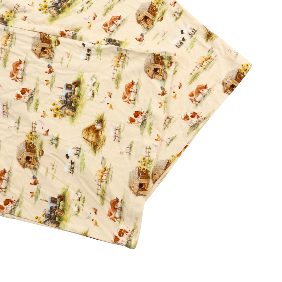 A folded quilted blanket with a cream-colored background and a detailed farm animal and cottage pattern, featuring brown, green, and orange illustrations of cows, chickens, and rustic cabins.