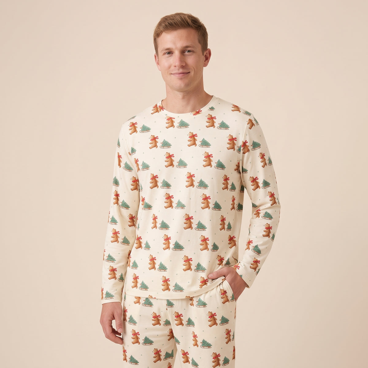 A man wearing a long-sleeve pajama shirt and matching pants with an all-over Christmas print of brown bears and green pine trees on a cream background, set against a solid beige studio backdrop.