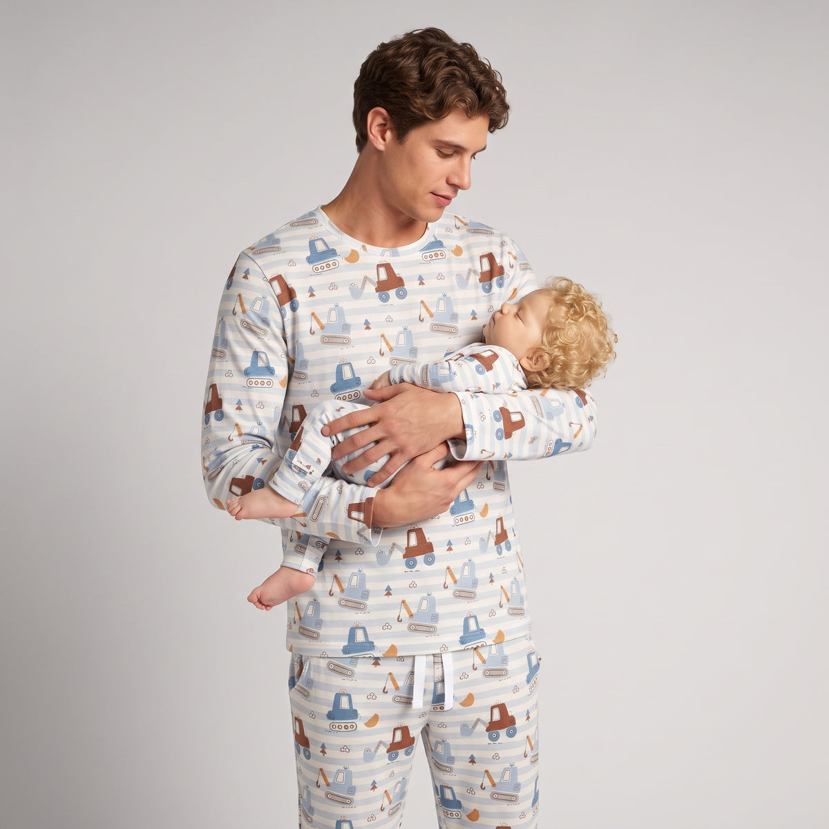 A man wearing a matching long-sleeve pajama set with a baby carrier print, holding a baby against his chest, photographed against a light gray studio background.