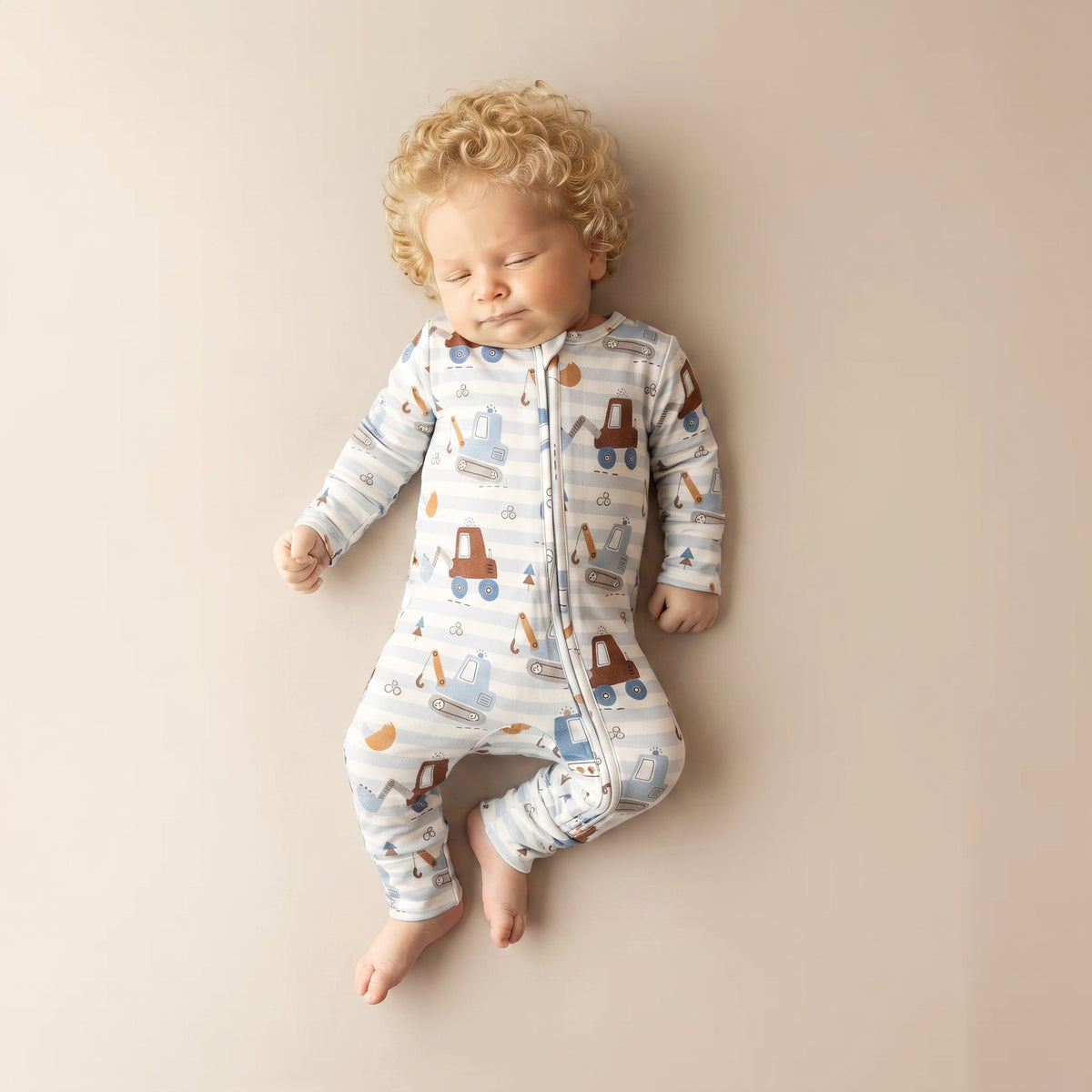 A sleeping baby with curly blond hair wearing a long-sleeve zip-front sleeper with a blue, white, and brown construction truck pattern, lying on a beige background.