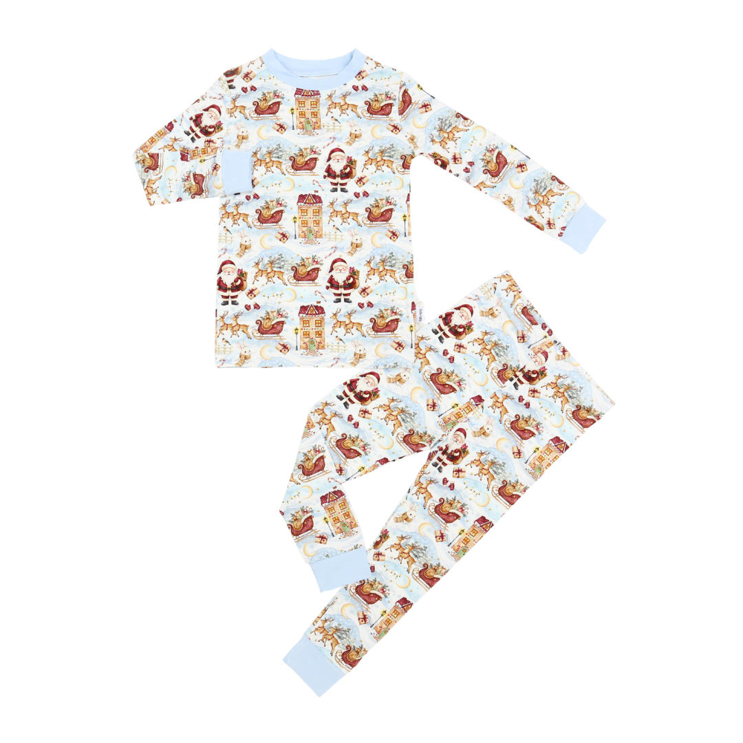 A two-piece children's pajama set featuring a long-sleeve top and matching pants with an all-over festive Christmas print. The print includes Santa Claus, reindeer, sleighs, gifts, and snowflakes on a white background, finished with light blue ribbed cuffs and collar.