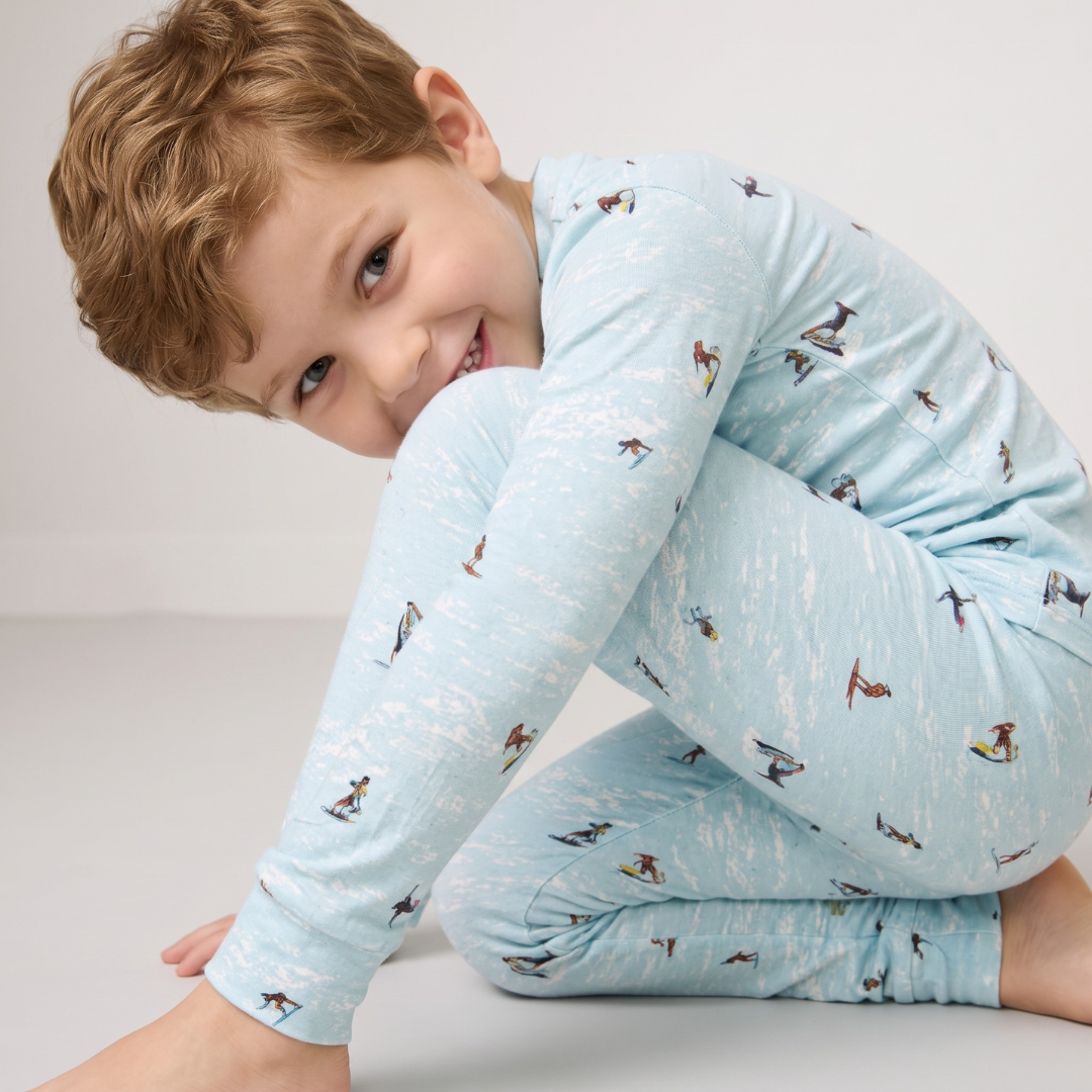 A young boy with curly light brown hair wearing a light blue long-sleeve pajama set with a small penguin print, posing on a light gray studio floor.