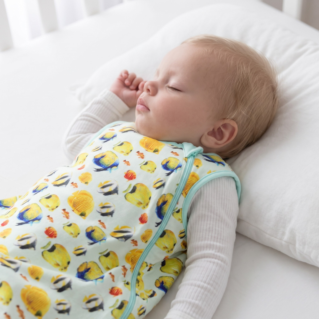 Baby sleeping on a white pillow wearing a colorful duck patterned outfit.