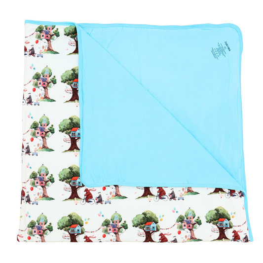 Children's blanket with tree house pattern on one side and solid blue color on the other, displayed against a white background.