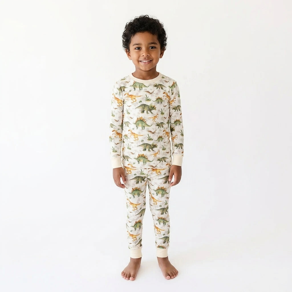 A young child standing against a plain white studio background, wearing a long-sleeve pajama set with an allover dinosaur print in green, orange, and brown on an off-white base.
