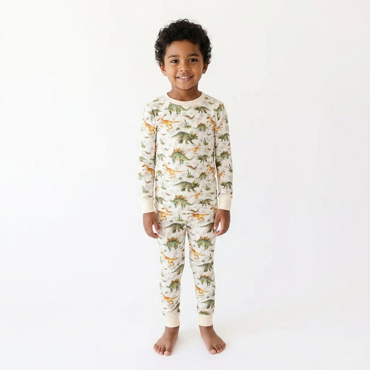 A young child standing against a plain white studio background, wearing a long-sleeve pajama set with an allover dinosaur print in green, orange, and brown on an off-white base.