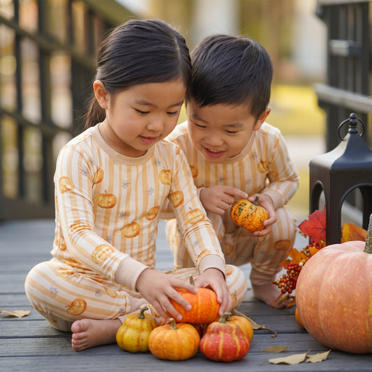 Two children sitting on a wooden deck wearing matching pajamas with a pumpkin and stripe pattern, surrounded by small pumpkins and autumn decorations.