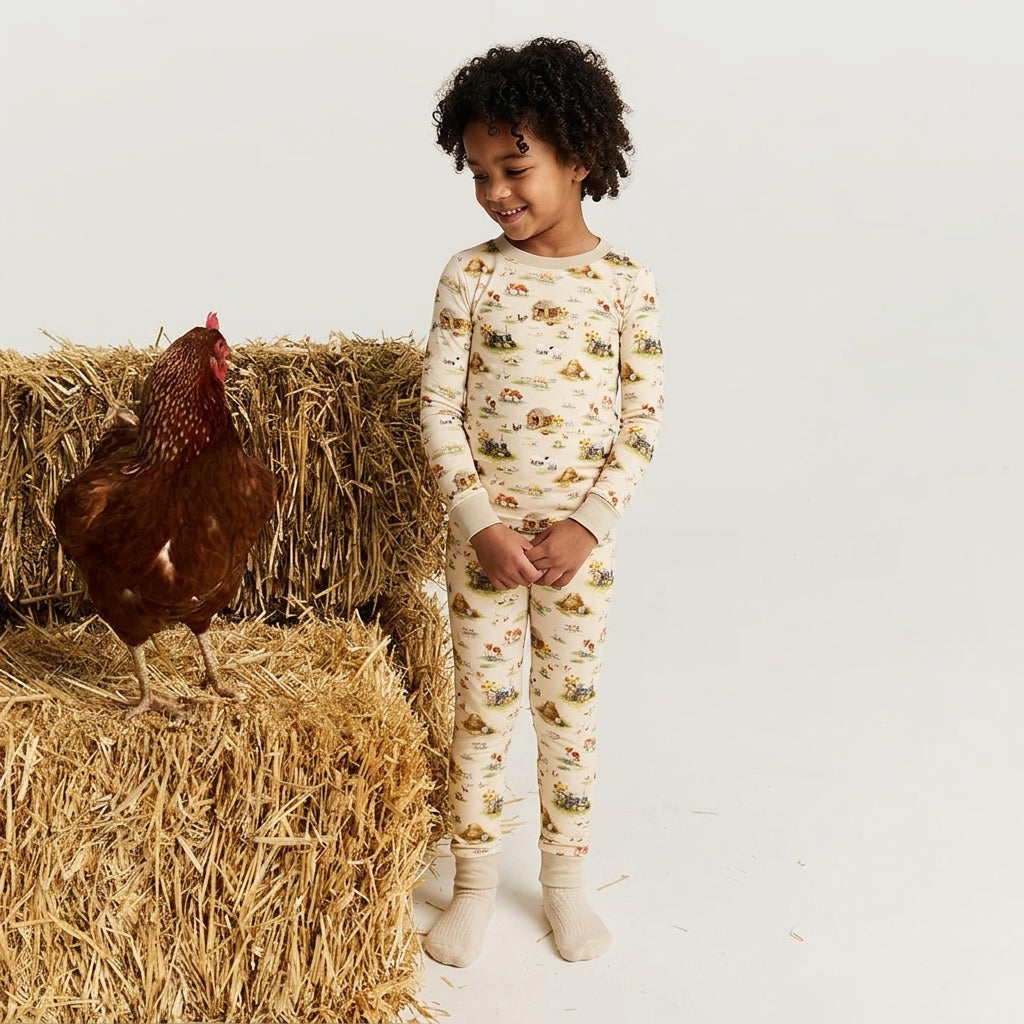 A young child wearing a cream-colored long-sleeve pajama set with a small farm animal print stands next to a brown rooster on a bale of hay against a plain white studio background.