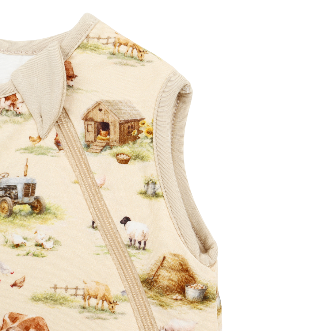 Close-up of a baby sleep sack featuring a cream-colored background with a detailed farm animal print including cows, sheep, a tractor, and a chicken coop, finished with a zippered front.