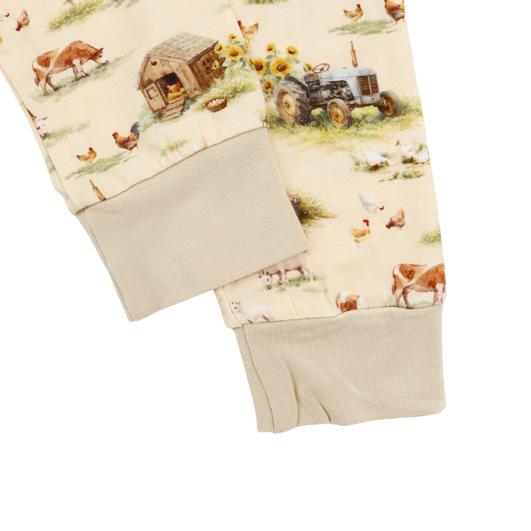 Close-up of a pair of fleece pajama pants with a cream-colored background and a detailed farm print featuring a tractor, barn, chickens, cows, and sunflowers. The pants have ribbed beige cuffs.